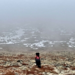 Revna sits on a mountainside among rocks and red tundra looking out over a valley as if she is contemplating life.